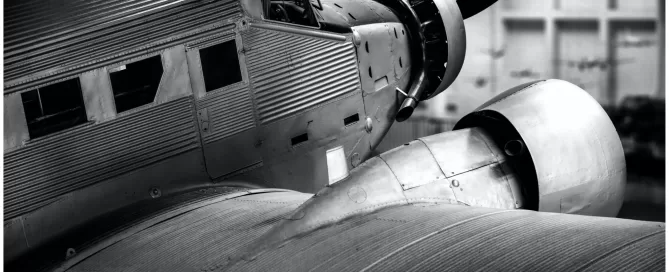 Black and white image of a vintage propeller plane viewed from the side, showing details of its corrugated metal body and engine.
