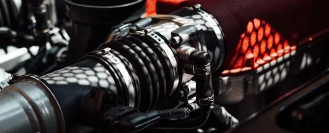 Close-up of a car engine with red covers, featuring intricate metallic components and a mesh pattern under sunlight.