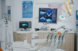 A dental clinic with an examination chair, overhead light, digital X-rays on monitors, dental tools, and a lab coat on a hanger is meticulously designed to meet industry standards.