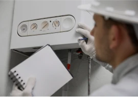 A man in a hard hat and gloves inspects a boiler unit while holding a notepad and pen.