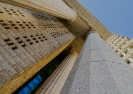 Low-angle view of a monumental building with tall stone columns and a textured facade against a clear blue sky.