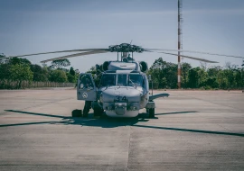 A helicopter sits on a tarmac with its blades outstretched, epitomizing the marvels of aerospace engineering. A person is observed inside the open side door. Trees and a tall antenna can be seen in the background.