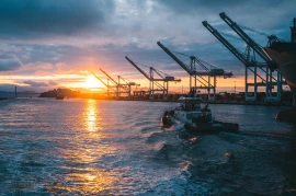 Sunset at a port with large cranes silhouetted against the sky. Industries humming with activity as a boat moves through the water, leaving a wake behind.