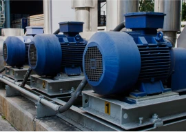 Three blue industrial electric motors are mounted on a concrete platform with metal pipes and conduits connected to them.