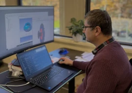 A man in glasses and a maroon sweater works at an office desk with a laptop and a large external monitor displaying a 3D model and code. Papers and a plant are on the windowsill behind him.
