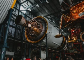 A large rocket engine is displayed indoors, with several people observing it from below. The surrounding structure is metallic and industrial.