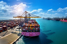 A large container ship docked at a busy port with cranes unloading colorful shipping containers, highlighting the vital industries under a sunny, partly cloudy sky.
