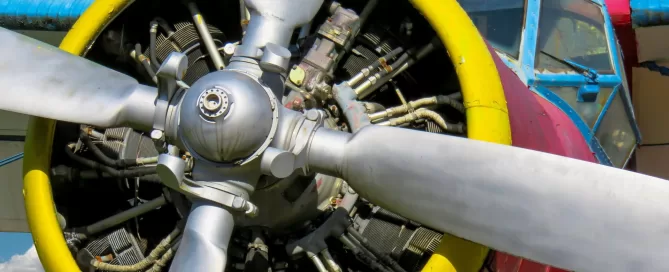 Close-up of an airplane's propeller and engine assembly showing metal blades, yellow engine rim, and red fuselage with a partly cloudy sky in the background.