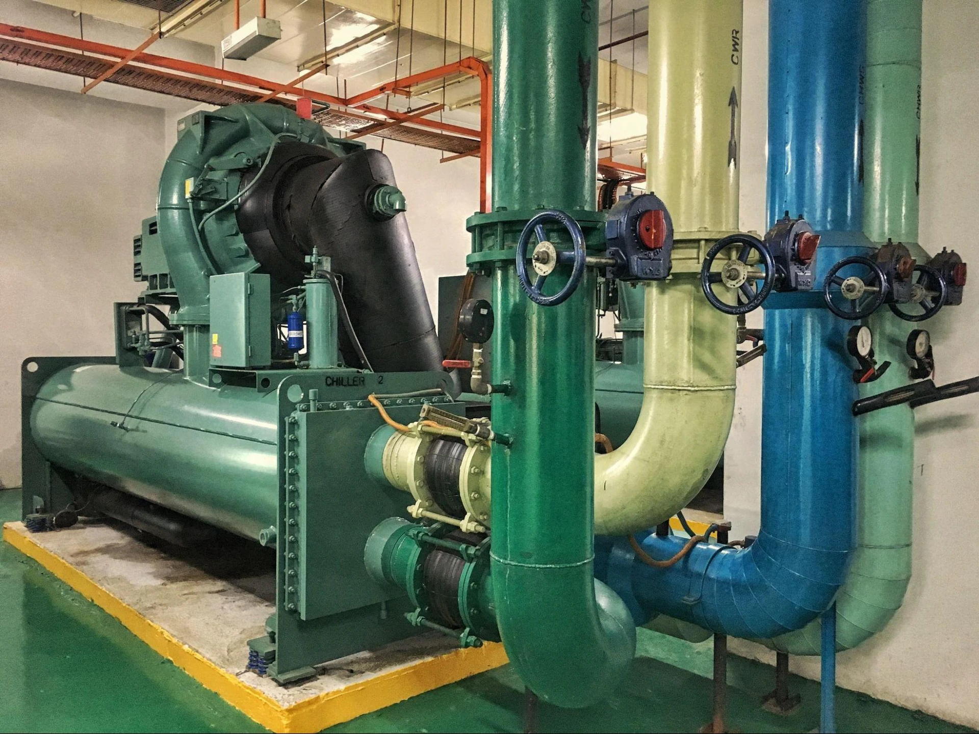 Industrial chiller system with large green and blue pipes, valves, and a motor in a mechanical room.