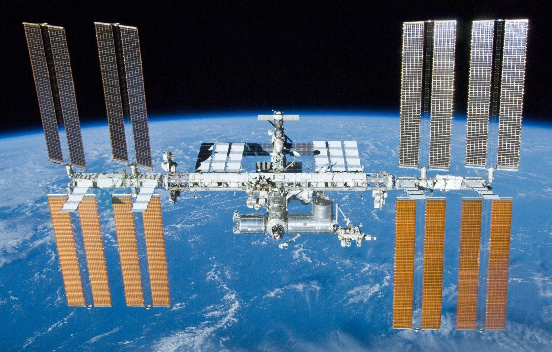 The International Space Station orbits above Earth with solar panels extended against a backdrop of space and the planet's curved horizon.