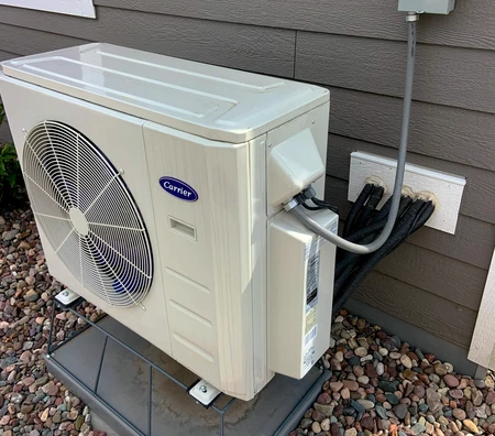 Outdoor air conditioning unit installed on a raised platform next to a building wall, with black cables running inside. Surrounded by rocks.