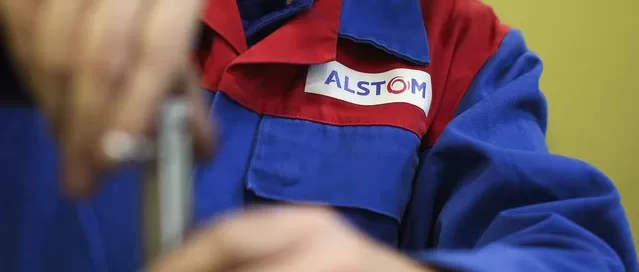 Person wearing a blue and red work uniform with an "Alstom" logo, holding a tool.