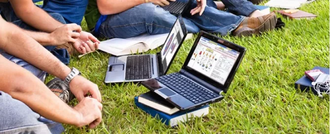 People sitting on grass with laptops and books, some writing in notebooks or working on devices.