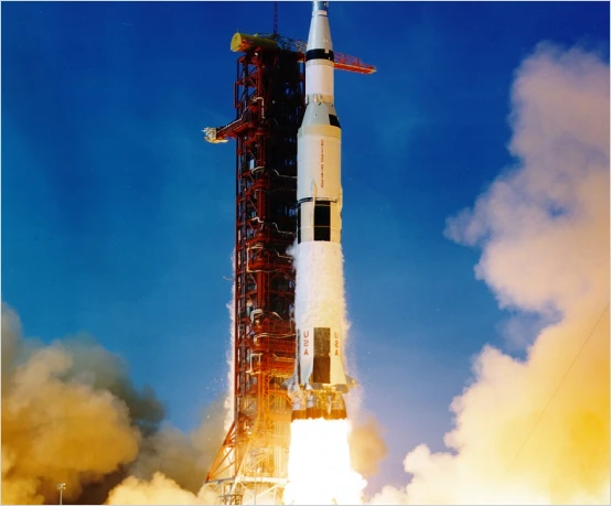 A Saturn V rocket, a marvel of technology, launches with bright flames and smoke against a clear blue sky.