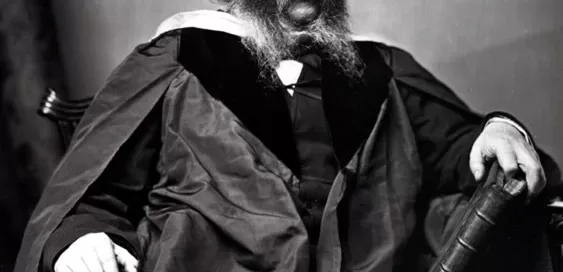 A seated man with long hair and a bushy beard wears a formal robe and holds a book.