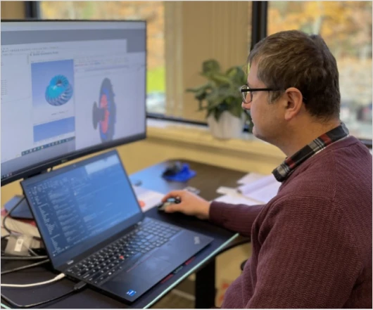 A person works at a desk with a laptop and a large monitor displaying technical diagrams related to turbomachinery development. A potted plant sits by the window, adding a touch of serenity to the workspace.