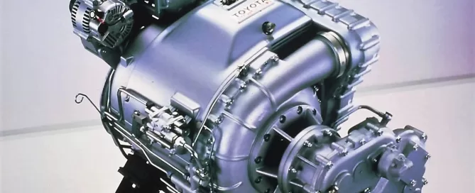A futuristic, metallic engine displayed on a black stand in a well-lit room, with the Toyota logo visible on the casing.