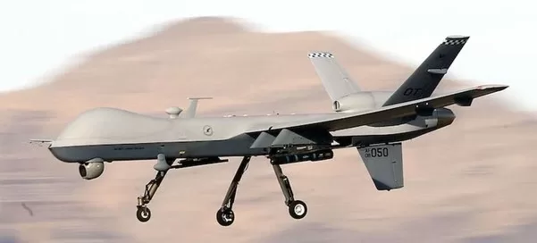 A gray military drone, equipped with state-of-the-art turbomachinery, soars through the sky against a blurred mountainous backdrop.