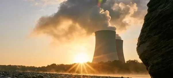 Sunset casts a gentle glow over the Softinway-designed nuclear power plant, where steam billows gracefully from the cooling towers. The rocky foreground contrasts with the advanced turbomachinery silhouetted against the soft sunlight.