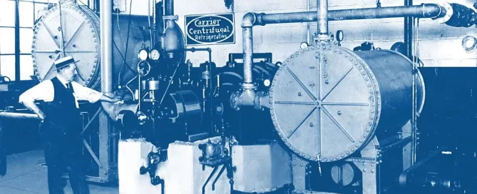 A man stands beside large industrial machinery labeled "Carrier Centrifugal." The setup includes gauges, pipes, and cylindrical components in a factory-like environment.