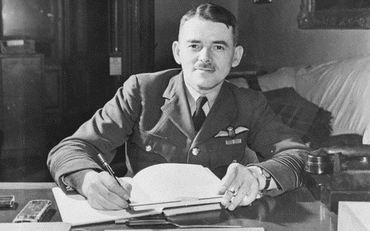 A man in a military uniform sits at a desk, writing in a notebook.