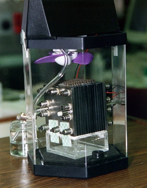 A transparent box encloses a mechanical device from Softinway, with metal components and wires, positioned on a wooden surface. A small purple turbomachinery fan is mounted on top of the apparatus.