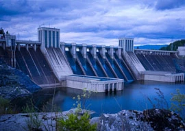 A large concrete dam with multiple spillways harnesses renewable energy technology, surrounded by water and a rocky area in the foreground under a cloudy sky.