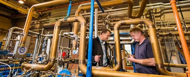 Two men stand among a network of pipes and machinery in an industrial setting, discussing a component.