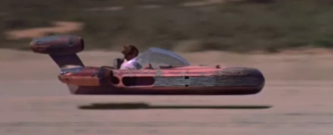 A person pilots a red and gray landspeeder, hovering above a sandy surface with blurred background.
