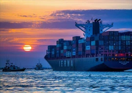 A large cargo ship with stacked containers, a marvel of transportation technology, sails on the ocean at sunset, accompanied by smaller boats.