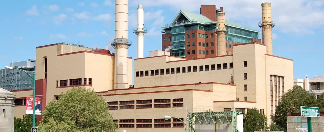 A large industrial building with multiple chimneys stands in an urban area. A tall, red brick building is visible in the background, and a metal fence lines the foreground.