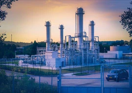 A gas processing facility, integral to power generation, stands with its tall structures and pipelines enclosed by a fence. A vehicle is parked nearby under the vibrant hues of a sunset sky.