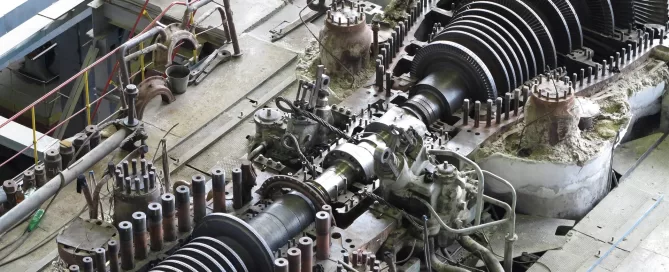 View of a disassembled industrial turbine with exposed rotors and numerous components, situated in a large maintenance area with tools and pipes visible in the background.