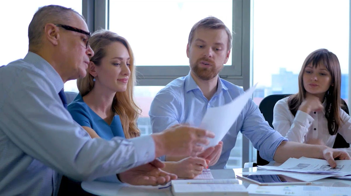 Four people sit around a table in an office setting, deep in discussion over fan design documents. A man points at the papers held by another person, emphasizing crucial details in their collaborative effort.