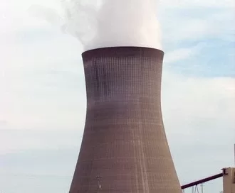 Cooling tower of a nuclear power plant releasing steam against a cloudy sky, surrounded by grass and industrial structures.