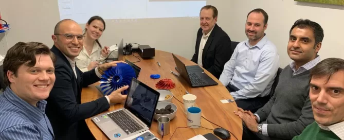 Seven people are seated around a table in a meeting room. They have laptops open, with projector slides on the wall. A blue 3D model and notebooks are on the table. A lion poster is on the wall.
