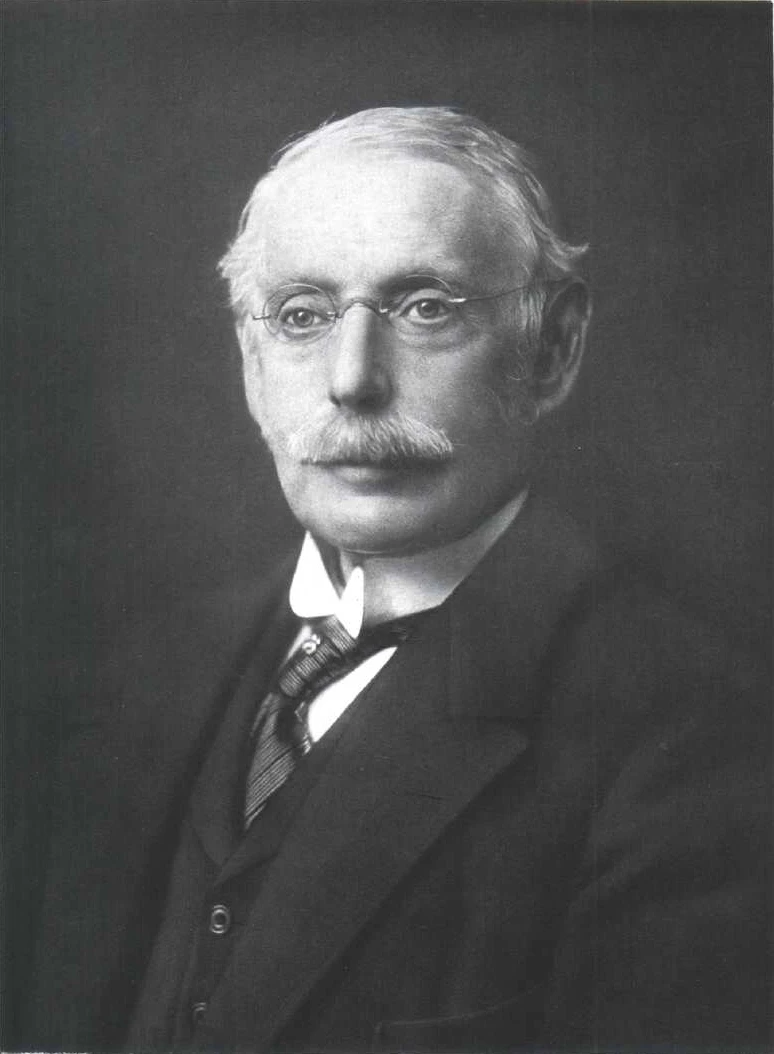 Black and white portrait of an older man with glasses, a mustache, and receding hairline wearing a suit and tie. Beige background.