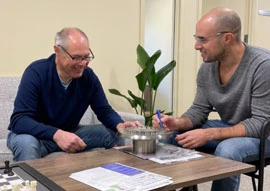 Tailored Solutions Two men sit at a table, smiling and deeply engaged in conversation about software customization. One holds a pen and paper, jotting down notes. A plant is in the background, adding a touch of nature to their discussion setting.