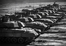 A line of tanks, symbols of government and defense, stand parked in a desert landscape under a cloudy sky, captured in black and white.