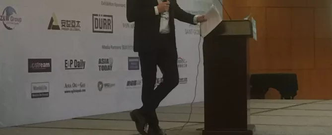 A person in business attire speaking at a podium on a stage with a backdrop of logos at a conference.