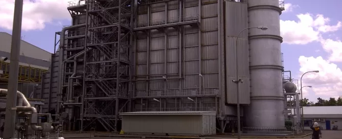 Industrial facility with large metal structures and a tall cylindrical tower. A person on a bicycle is visible in the foreground under a partly cloudy sky.