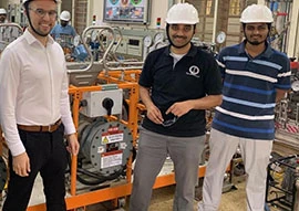 Three people in hard hats stand in an industrial aerospace setting near machinery, smiling at the camera.