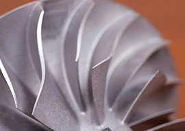 Close-up of a metallic turbine or fan blade, reminiscent of an advanced appliance, with a radial design featuring sharp, curved edges against a warm brown background.