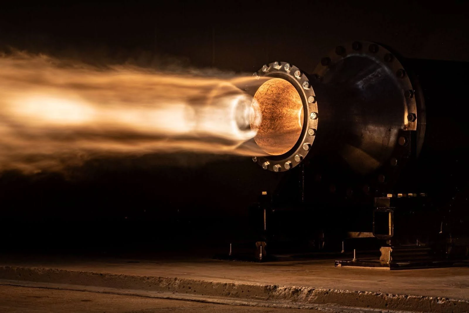 government-research-and-development-5 In a dimly lit industrial setting, a large engine or nozzle emits a bright, fiery exhaust plume horizontally, showcasing the cutting-edge advancements stemming from government research.