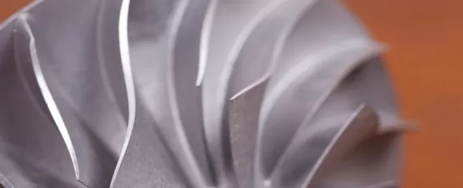 Close-up of a metallic turbine blade with curved, sharp edges against a blurred, brown background.