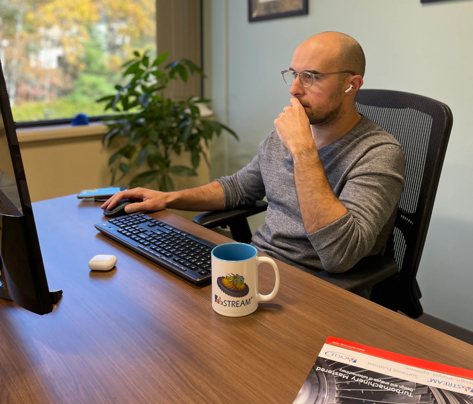 software-customization-1 A man with glasses focuses on software customization at his computer in an office, earbuds in place. A mug and notebook rest on the desk beside him. Behind, a large window lets light spill over a potted plant, adding to the serene workspace ambiance.