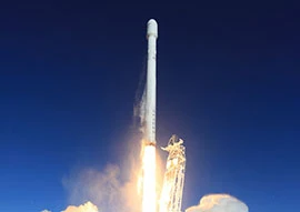 A rocket, showcasing the marvel of space technology, is launching into a clear blue sky with flames and smoke at its base.