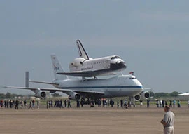 A marvel of space technology, the space shuttle piggybacks gracefully on a NASA Boeing 747, nestled on the runway as a fascinated crowd gathers at the airport.