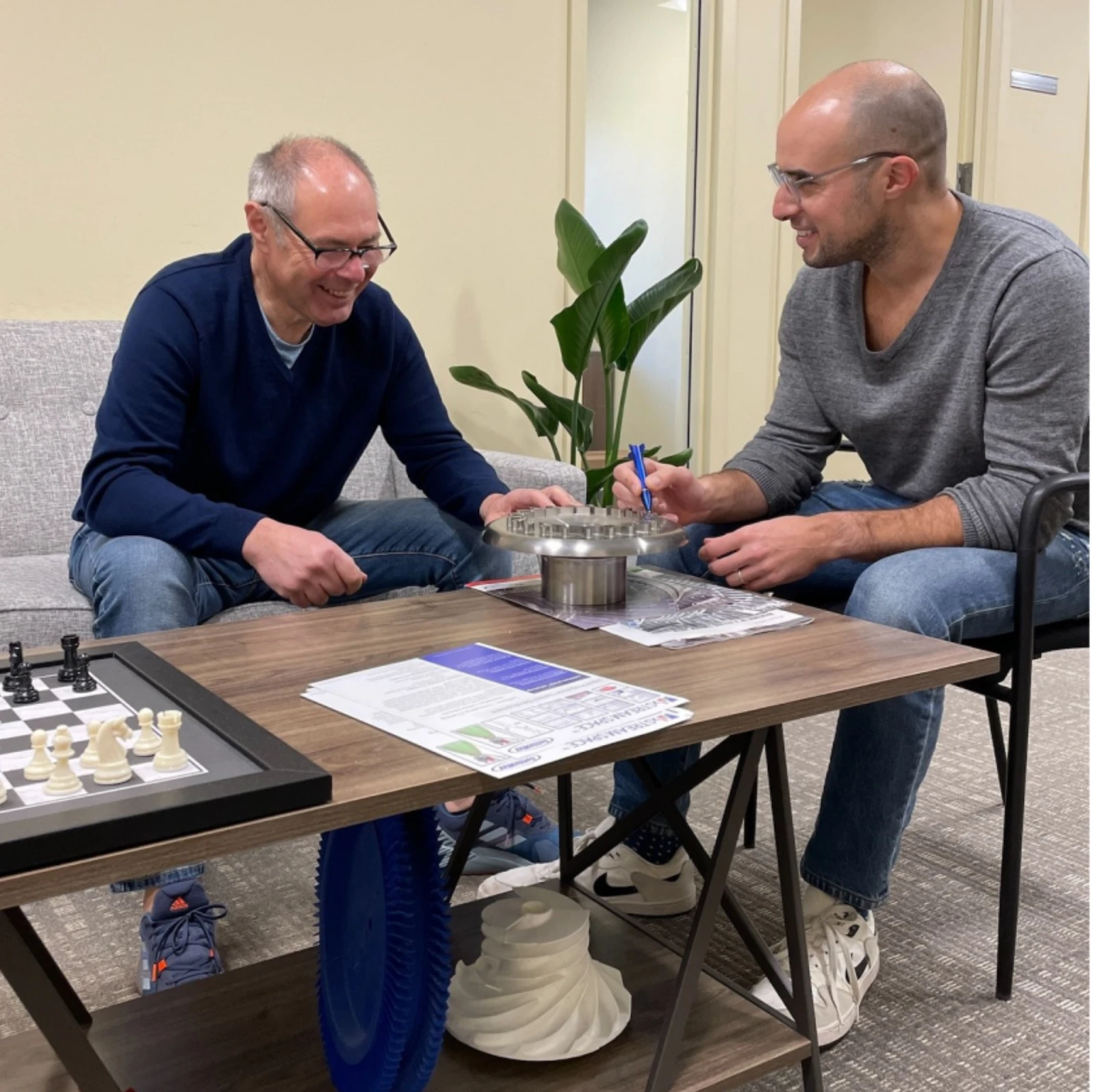 Two men sit at a table discussing a mechanical part, surrounded by documents and a chessboard. A plant adds a touch of green to their workspace as they dive into Thermal-Fluid System Modeling.