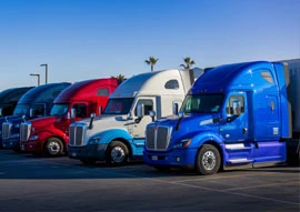 A row of colorful semi-trucks, showcasing the latest in transportation technology, is parked in a lot under a clear blue sky.
